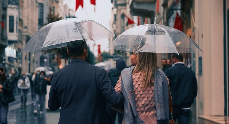 Couple walking in the rain with clear umbrellas on a bustling city street, seen from behind.