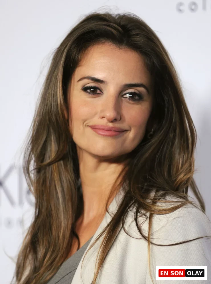 Penelope Cruz, esmerlere yakışan uzun, dalgalı, kahverengi saçları ve hafif karamel balyajlarıyla gülümseyerek kameraya bakıyor; bu görünüm, esmerler için ideal saç renkleri seçeneklerinden birini başarıyla sergiliyor.