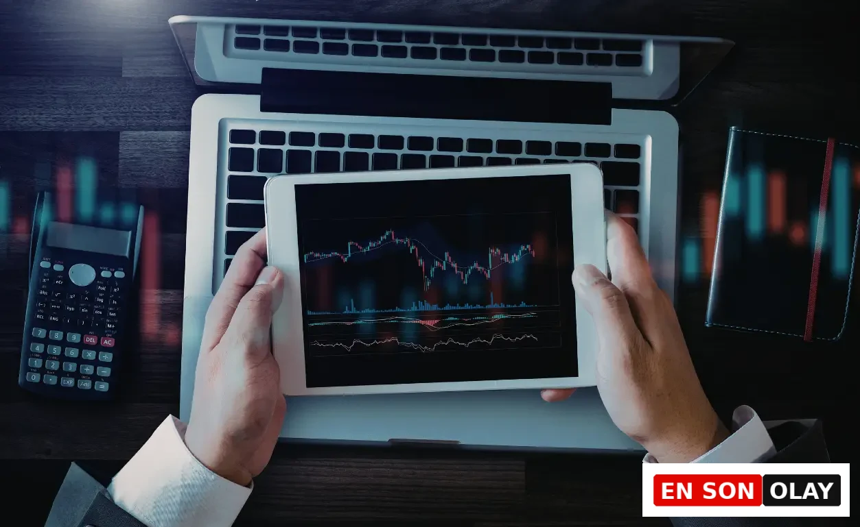 Bir iş profesyonelinin dizüstü bilgisayarın üzerinde finansal grafikler gösteren bir tableti iki eliyle tutarak, yanında bir hesap makinesi ve not defteriyle karmaşık piyasa verilerini dikkatle incelediği bu görsel, uzun süreli analiz ve ardışık kararların getirebileceği karar yorgunluğunu çağrıştırıyor.