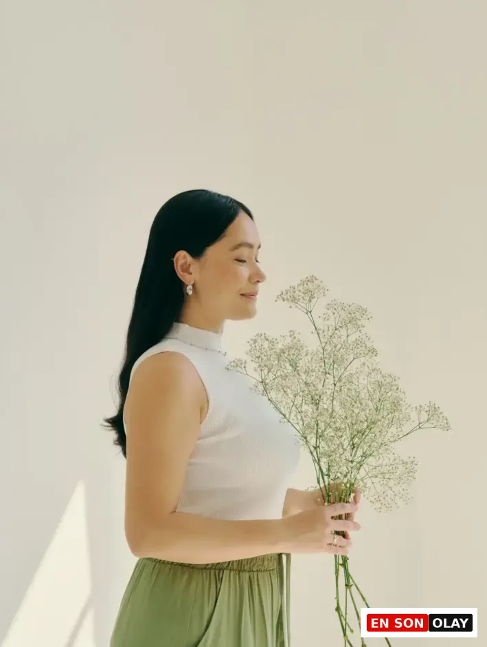 Güneş ışığında aromaterapi etkisiyle rahatlayan kadın; elinde beyaz çiçek buketi tutuyor, gözleri kapalı ve huzurlu bir şekilde nefes alıyor. Minimalist arka plan ve doğal ışıkla içsel dengeyi yansıtan görsel.