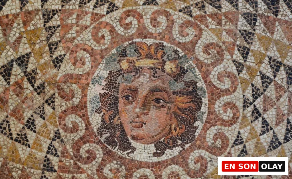 Antik bir mozaiğin merkezinde, yaprak ve sarmaşıklarla süslenmiş bir çelenk taşıyan, kahverengi tonlarında bukleli saçlara ve altın rengi küpeye sahip genç bir yüz, ayrıntılı mozaik taşlarıyla özenle işlenmiş; etrafı kahverengi ve beyaz sarmal desenlerle, dış kısımları ise üçgen geometrik motiflerle çevrilidir.