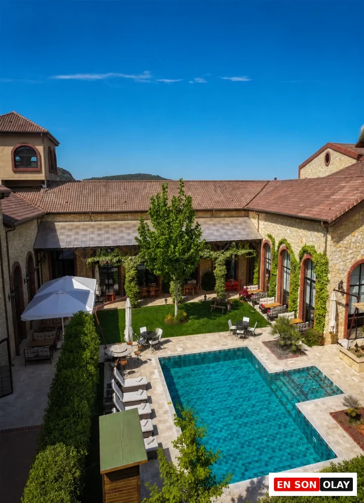 Çanakkale'de bulunan Bengodi Boutique Hotel'in kuşbakışı görüntüsünde, kiremit çatılı taş yapılarla çevrili yemyeşil bir bahçe içerisinde, güneşli mavi gökyüzü altında berrak mavi mozaik karolarıyla dikkat çeken bir açık yüzme havuzu ve etrafında şezlonglar ile zarif dış mekan oturma alanları görülüyor.