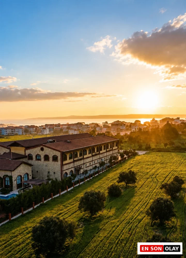 Gün batımının altın rengi ışıkları altında, Bengodi Boutique Otel'in kahverengi çatılı, taş detaylı binası, önündeki denize uzanan izli yeşil tarlalar ve uzaktaki sahil kasabasıyla birlikte huzurlu bir manzarayı gözler önüne seriyor.