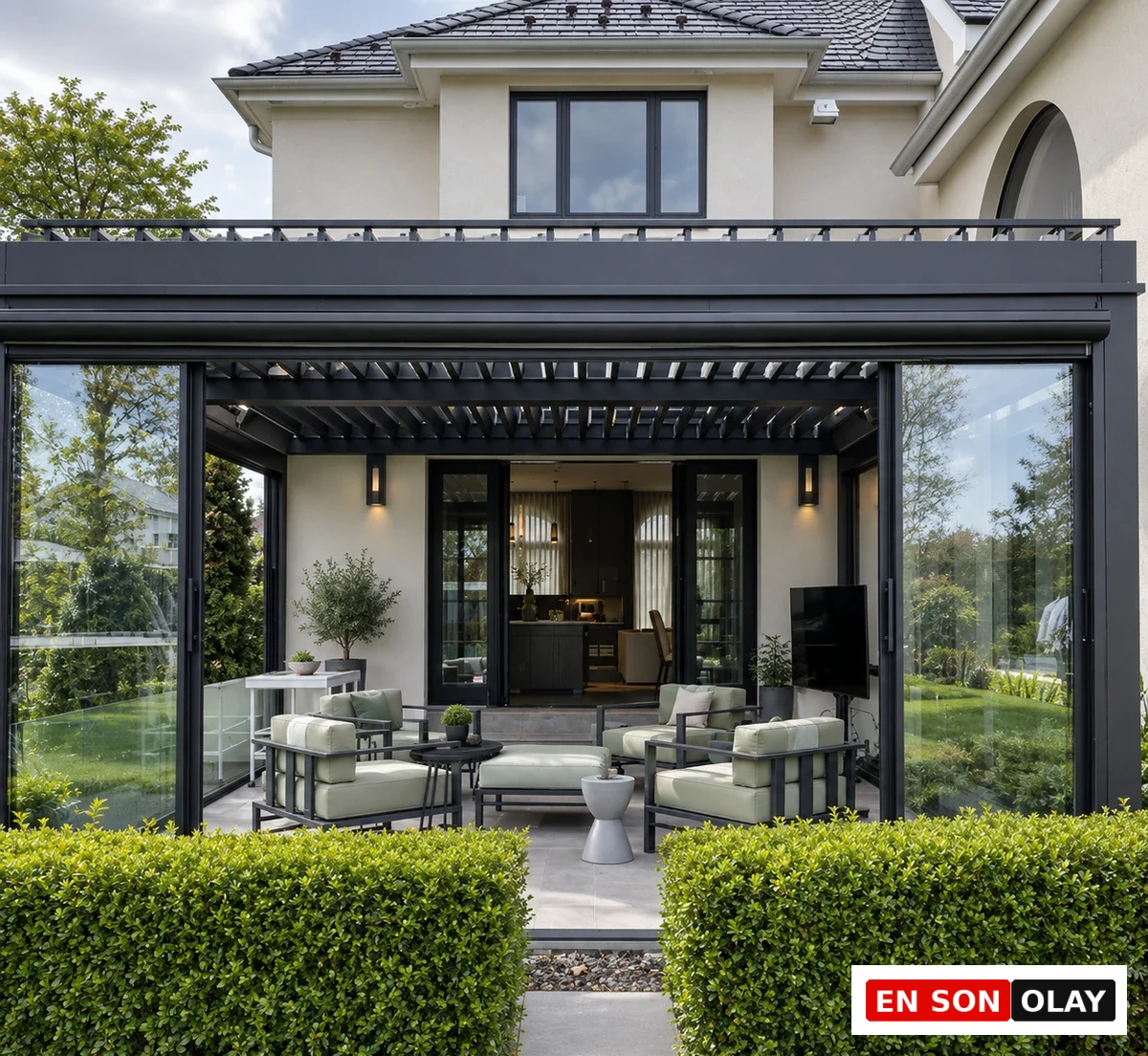 Büyük cam paneller ve modern açılır kapanır çatı sistemiyle tasarlanmış, krem rengi bir evin bahçesine entegre edilmiş şık bir kış bahçesi; içinde açık renk minderli modern dış mekan mobilyaları, bitkiler ve yeşil çitlerle çevrili geniş bir yaşam alanı sunuyor.