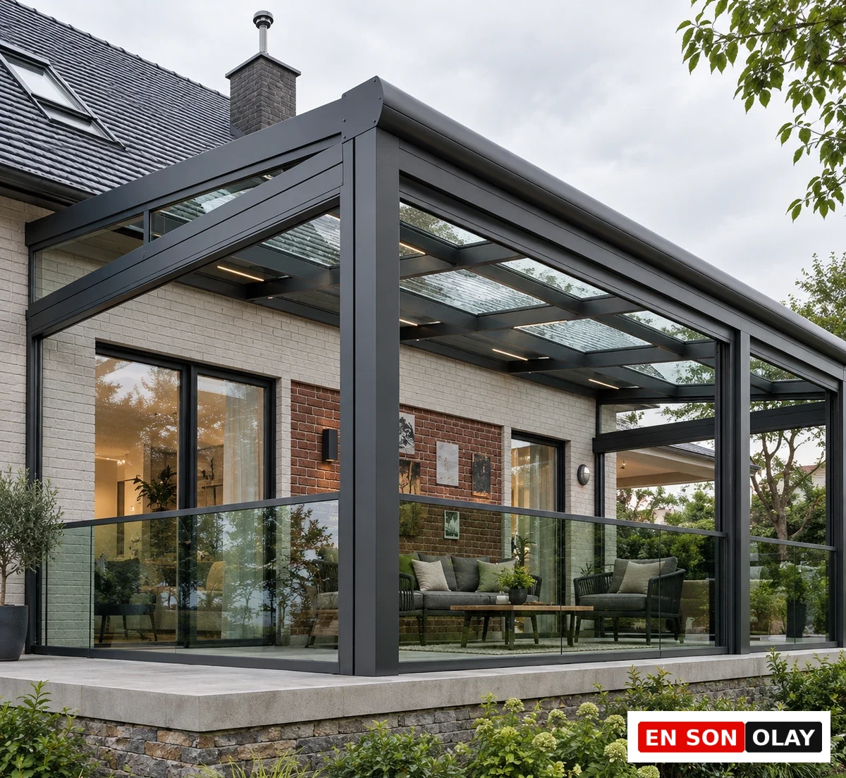 Gri metal profilli ve cam tavanlı modern bir kış bahçesi, dışarıda yeşil bitkilerle çevrili bir bahçenin önünde, açık renkli tuğla duvarlı bir villaya şık bir şekilde entegre edilmiş olup, ısı yalıtımlı camlarıyla konforlu bir dış yaşam alanı sunmaktadır.