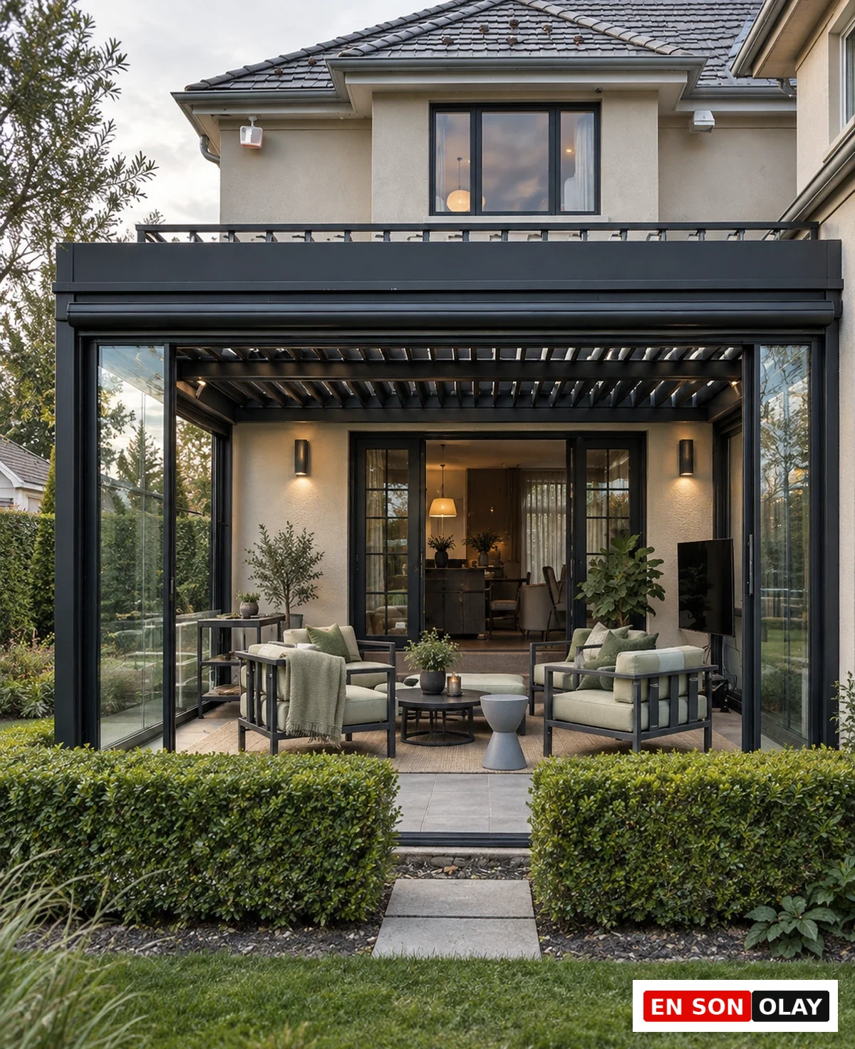 Villa cephesine entegre edilmiş, modern tasarımlı, koyu renkli bir rolling roof sistemi ve cam panellerle çevrili şık bir kış bahçesi modeli görülüyor; bu ferah alanda açık yeşil minderli konforlu oturma grupları, orta sehpa ve bitkiler yer alırken, dışarıda yemyeşil bir bahçe ve düzenli çitler estetik bir görünüm sunuyor.