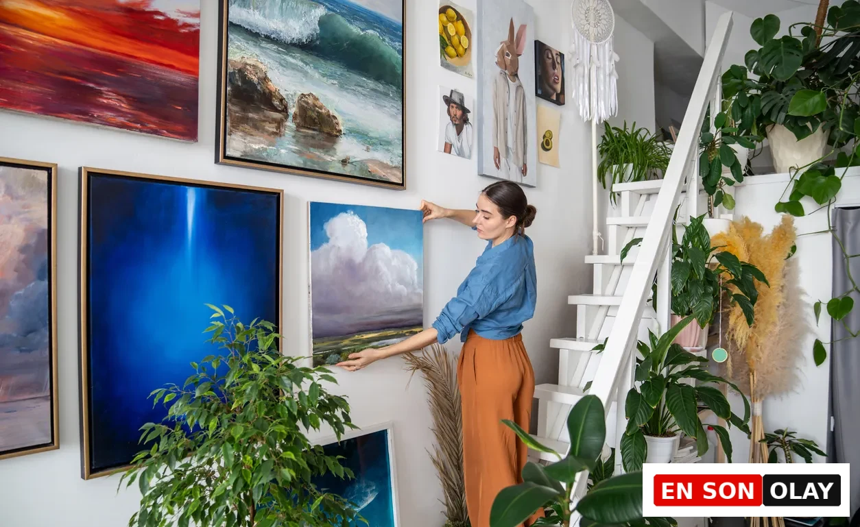 Bir kadın, ev dekorasyonu kapsamında, birçok farklı sanat eseriyle dolu beyaz bir duvara içinde bulutlu bir manzara olan çerçeveli bir tabloyu özenle asıyor; çevresi merdiven ve yemyeşil bitkilerle dolu, sanatsal bir yaşam alanı.