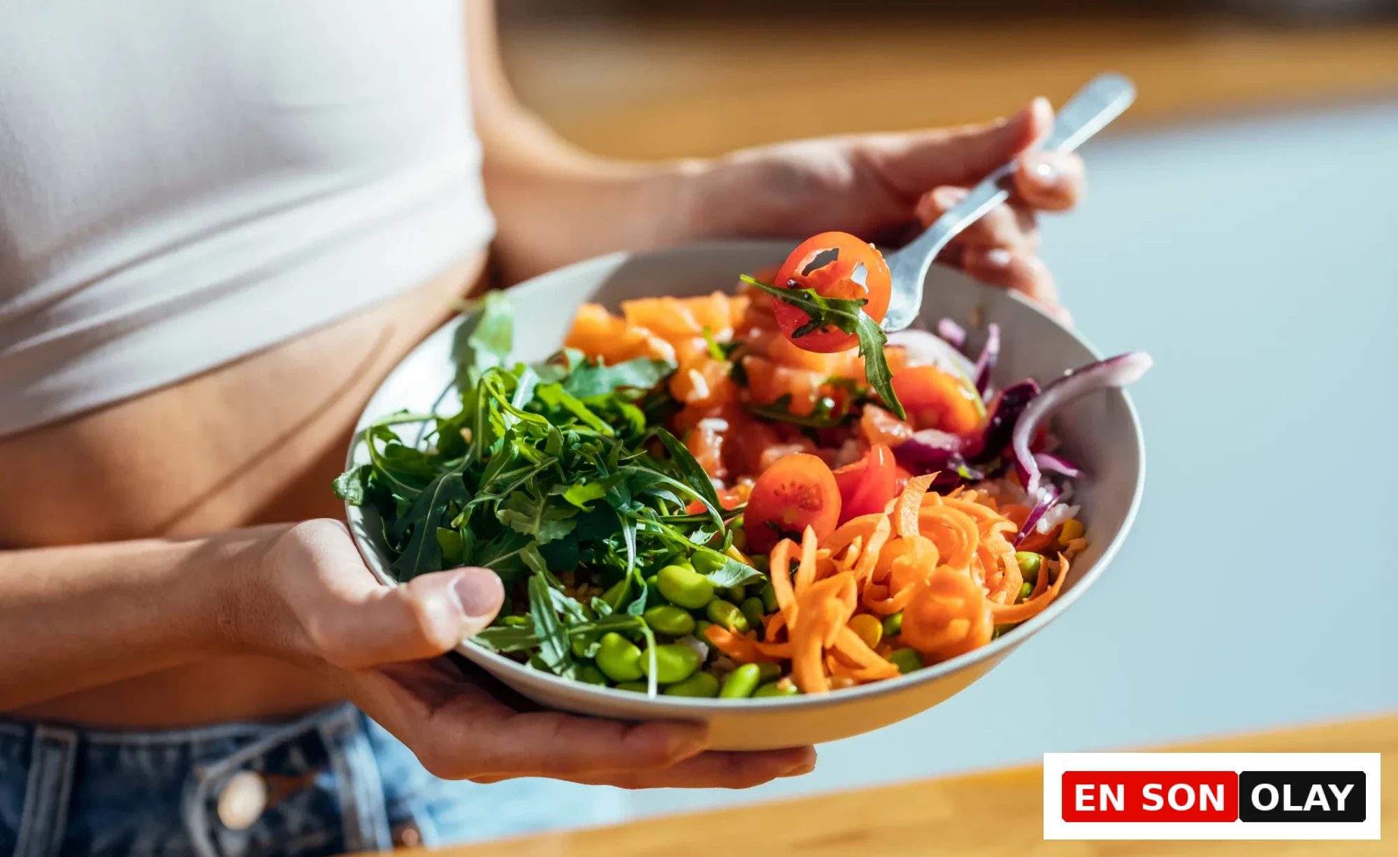 Beyaz bir atlet ve kot pantolon giyen bir kişi, taze roka, kiraz domates, rendelenmiş havuç, edamame ve kırmızı soğan içeren rengarenk, sağlıklı bir salata kasesini tutuyor, çatalında bir parça domates ve roka var, bu görüntü Başak burcu için önerilen Nisan ayı detoks enerjisi ve sağlık temasına uygun beslenmeyi vurguluyor.