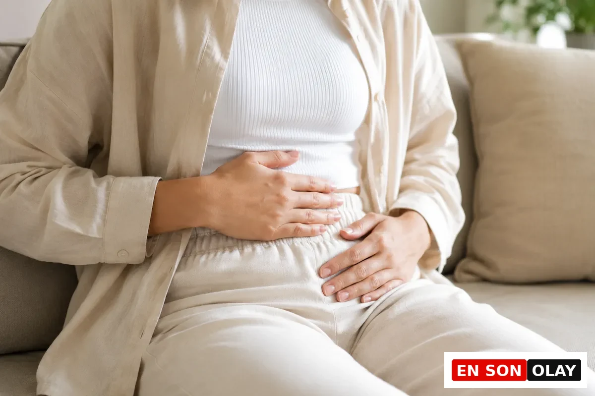Açık renk keten bir gömlek, atlet ve pantolon giyen bir kişi, ev ortamında bir koltukta otururken karın bölgesindeki rahatsızlık veya ağrıyı ifade etmek için ellerini nazikçe karnına bastırıyor, bu durum besin zehirlenmesi belirtilerinden biri olan mide sorunlarına işaret ediyor.