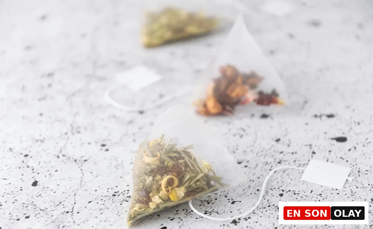 Besin zehirlenmesine iyi gelen çaylar konusuyla ilgili olarak, farklı kurutulmuş bitki ve meyve karışımları içeren, ip ve etiketiyle birlikte gri zemin üzerinde duran üç adet üçgen formda bitki çayı poşetinin yakın çekim görseli.
