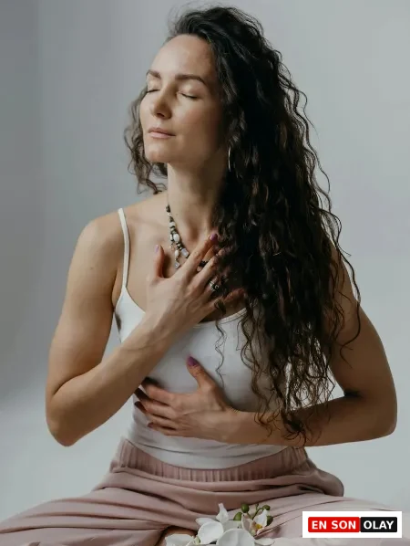 Uzun kıvırcık saçlı, beyaz bir atlet ve pembe pantolon giyen kadın, gözleri kapalı, elleri göğsünde meditasyon pozisyonunda bağdaş kurmuş olarak derin bir içsel odaklanma ve huzur içinde görünüyor; önündeki zarif beyaz orkidelerle birlikte bu görüntü, Theta Healing'in temelini oluşturan derin gevşeme ve bilinçaltı inanç çalışmasını doğal bir şekilde yansıtıyor.