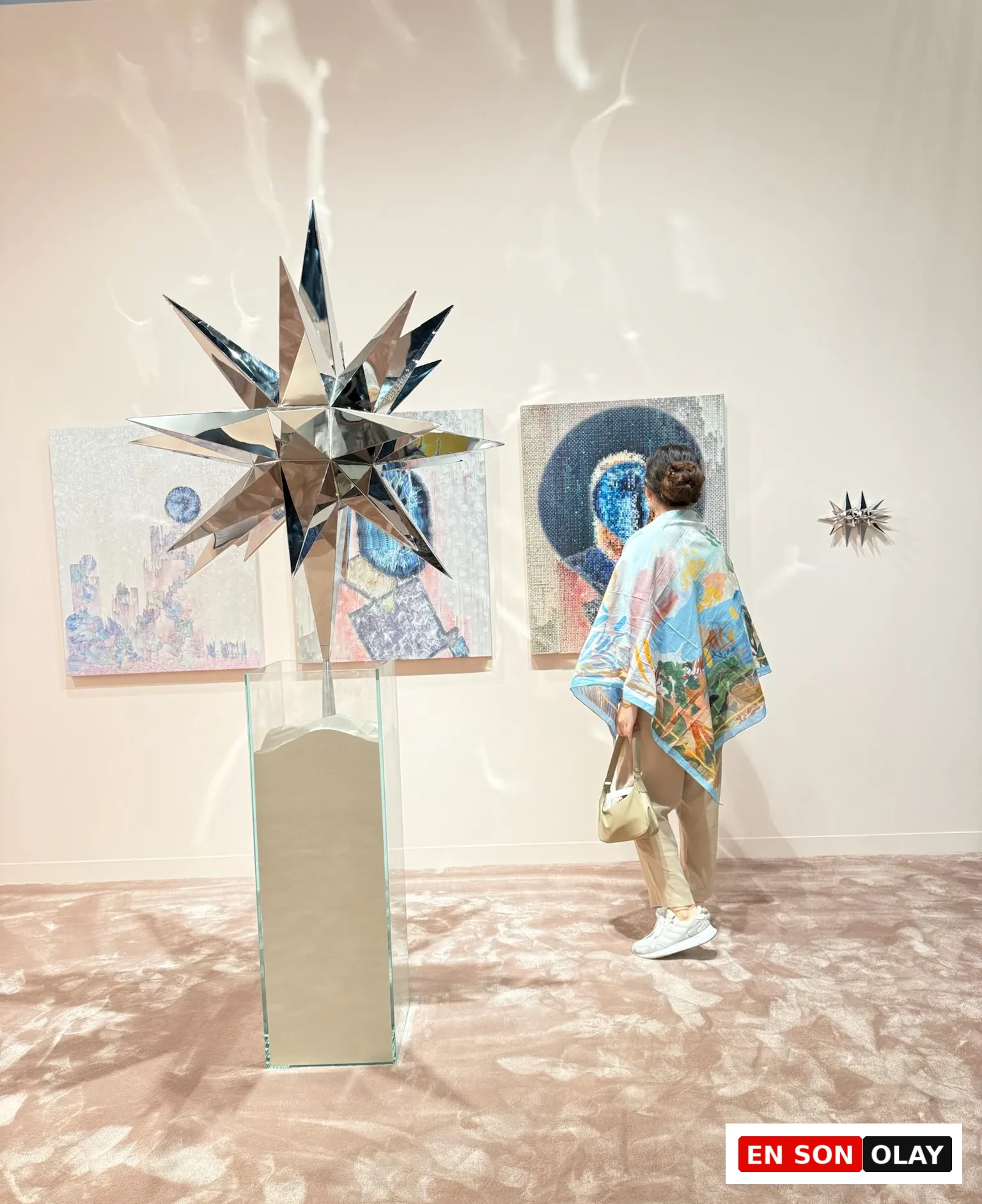 Abu Dhabi Art’ta ayna yüzeyli yıldız formunda bir heykel ve arka planda çağdaş resimlerin yer aldığı sergi alanını inceleyen ziyaretçi