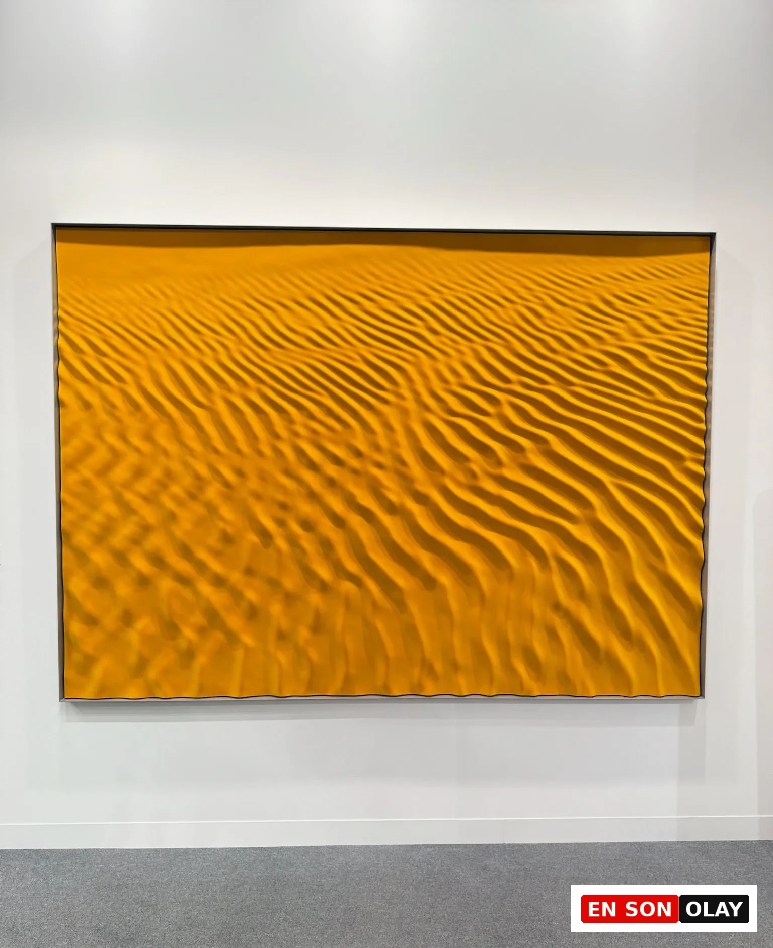 Abu Dhabi Art’ta sarı tonların hâkim olduğu, dalga dokulu soyut bir resmin sergilendiği galeri duvarı