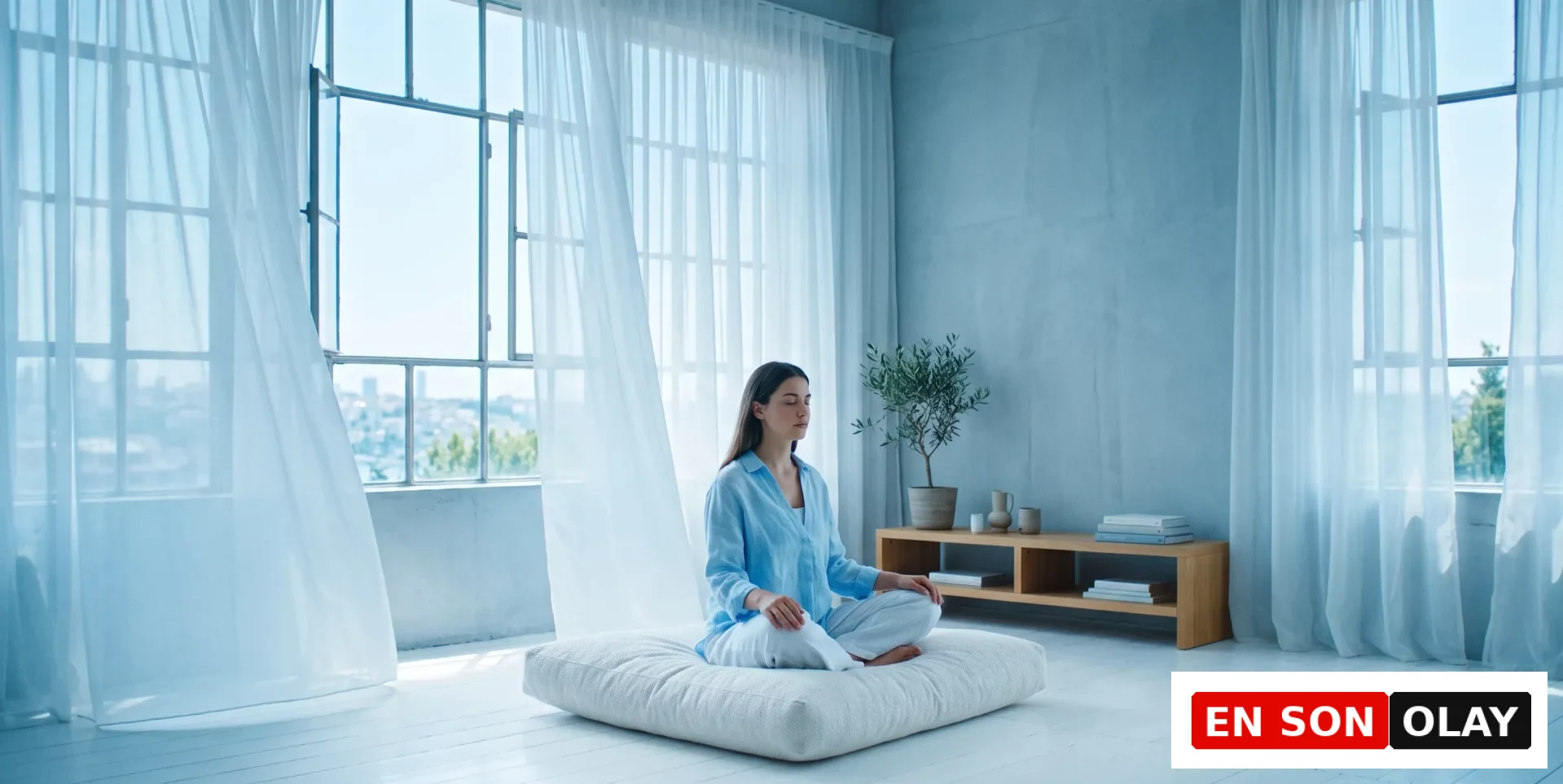 Ortada bağdaş kurmuş, gözleri kapalı, açık mavi gömlekli bir kadın, ferah ve aydınlık bir odada büyük pencerenin önündeki beyaz minder üzerinde meditasyon yaparak huzur bulmaya çalışıyor; hafifçe dalgalanan şeffaf perdeler ve odanın sakin atmosferi, stres yönetimi için ideal bir ortam sunuyor.
