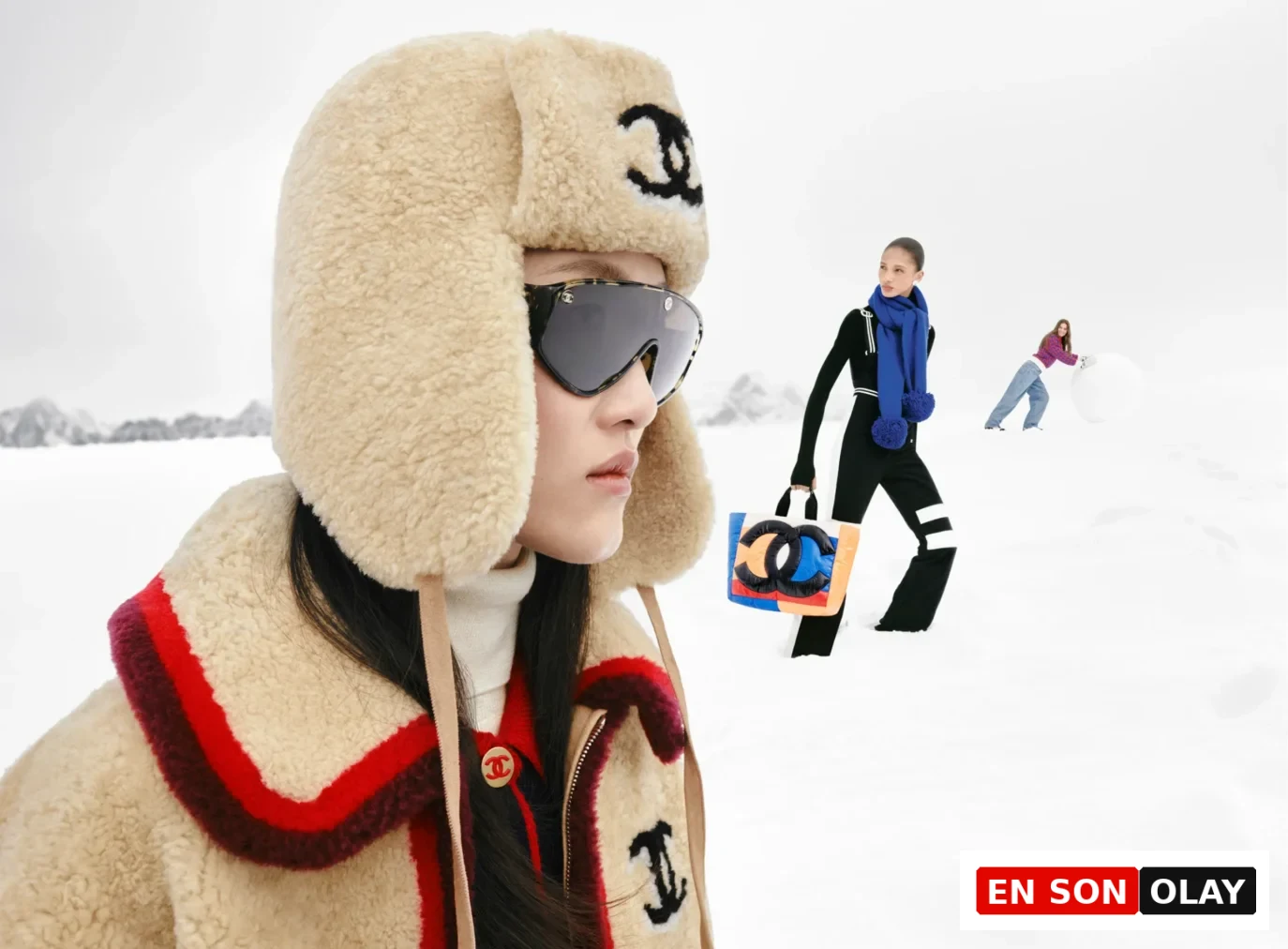 Ön planda kabarık bej bir kulaklıklı şapka ve büyük güneş gözlükleri takan model, karlı Megève manzarasında, geride renkli Chanel logolu bir çanta taşıyan başka bir model ve kar topu yuvarlayan bir kadınla, Chanel Coco Neige 2026'nın retro kayak modasına getirdiği sportif ve neşeli havayı gözler önüne seriyor.