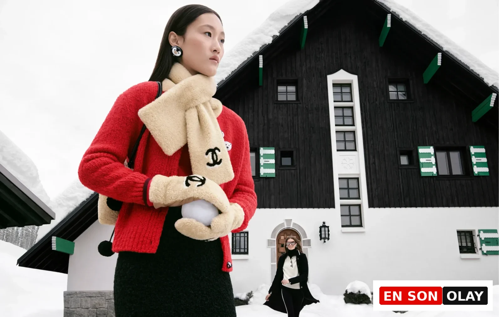 Karlı bir dağ evinin önünde, kırmızı hırkası, üzerindeki Chanel logosuyla bej renkli polar atkısı ve eldivenleriyle bembeyaz bir kar topu tutan model, Chanel Coco Neige koleksiyonunun retro kayak modasına getirdiği rahat ve şık kış stilini yansıtıyor.