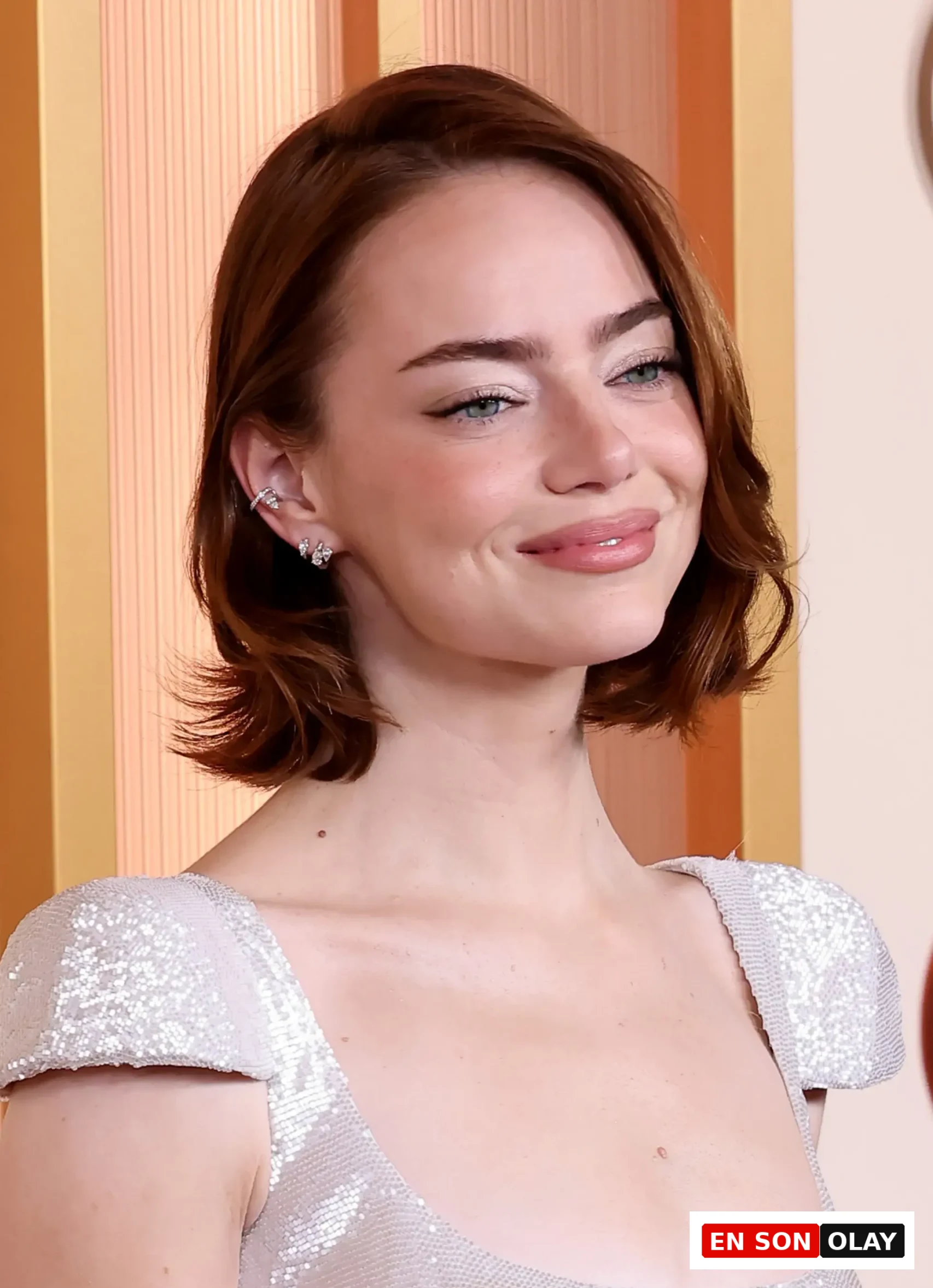 Oscar ödül töreni veya benzer bir etkinlikte, gümüş rengi pırıltılı, kap kollu bir elbiseyle hafifçe gülümseyen Emma Stone'un yakın çekim portresi; kızıl kısa saçları, mavi-yeşil gözleri ve parlak dudak makyajı dikkat çekiyor, sol kulağında birden fazla zarif küpe görülüyor.