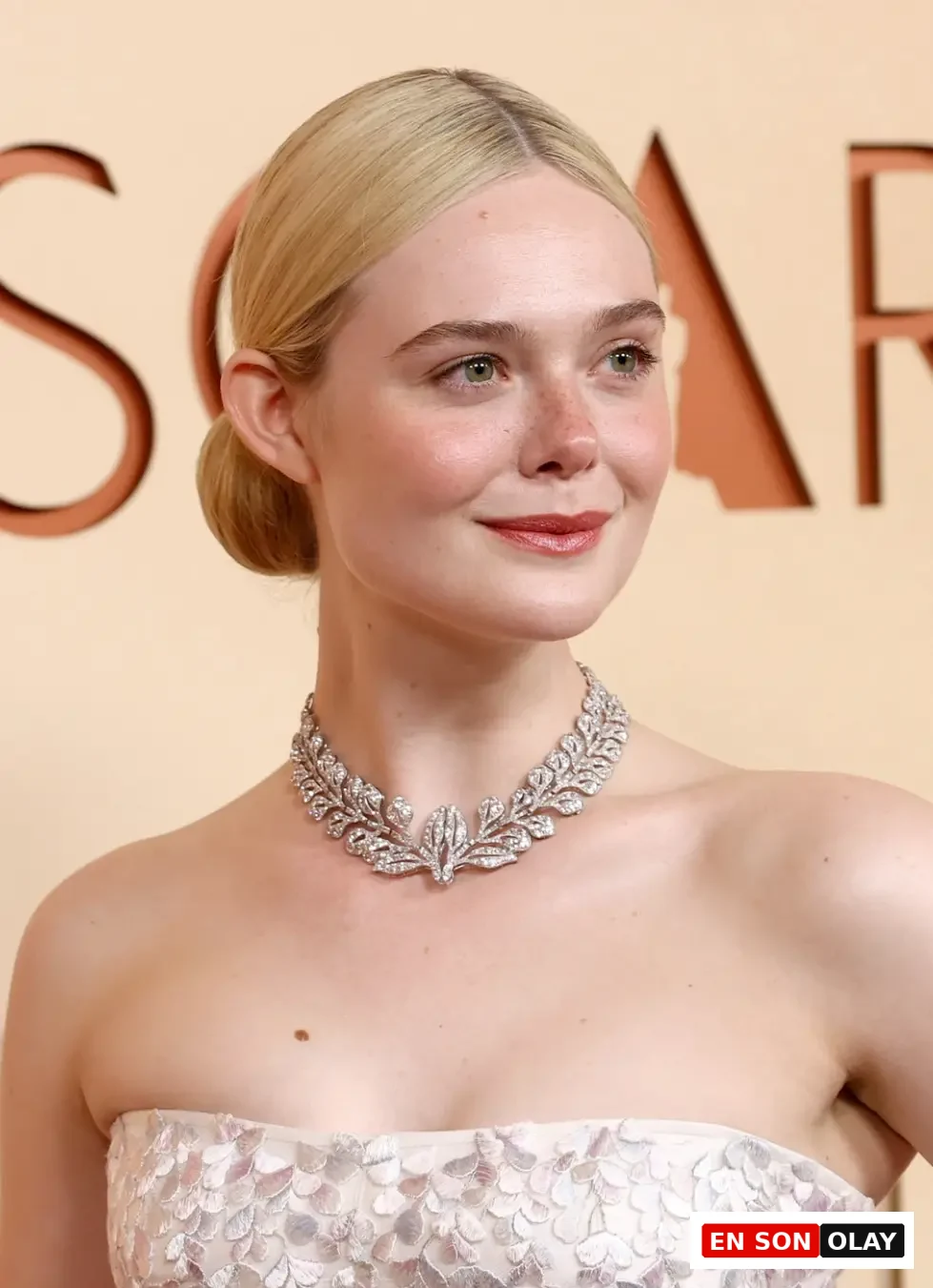 Elle Fanning, sarı saçları arkadan topuz yapılmış, ten rengi ve çilli yüzünde hafif bir gülümsemeyle, Oscar yazısının önünde, straplez, gümüşi yaprak işlemeli beyaz elbisesi ve göz alıcı yaprak desenli pırlanta kolyesiyle dikkat çekiyor.