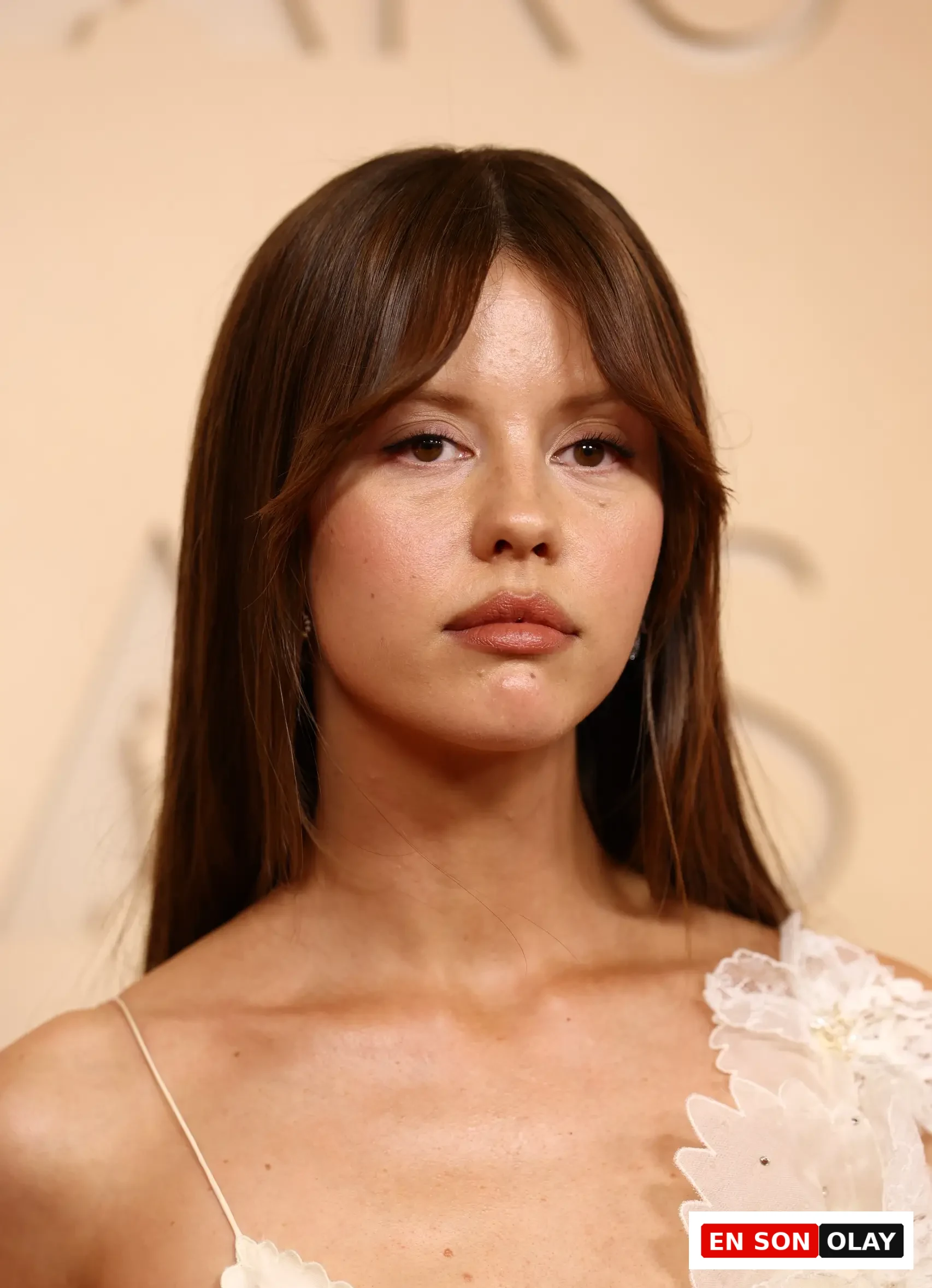Mia Goth, uzun kahverengi saçları ve alnına düşen perçemleriyle, hafif makyajlı yüzü ve dikkat çekici kahverengi gözleriyle kameraya doğrudan bakarken, zarif fildişi rengi, omuzunda işlemeli dantel detaylara sahip elbisesiyle 2026 Oscar töreniyle ilgili bir etkinlikte sonbahar cilt tonu ile görülüyor.
