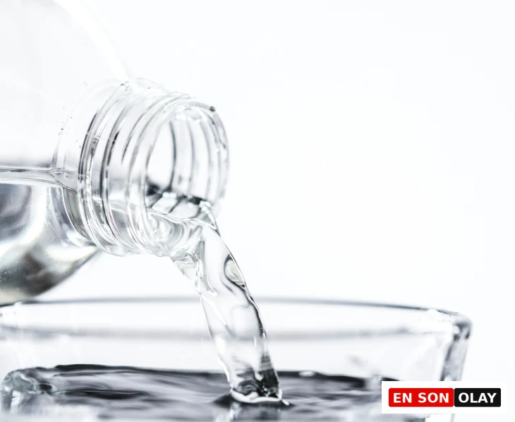 Beyaz arka plan önünde, şeffaf plastik bir şişeden şeffaf bir bardağa berrak su dökülürken, bu görsel kaliteli uykuya ulaşmak ve daha iyi dinlenmek için gün içinde dengeli su tüketiminin ve uyku hijyeninin kritik önemini hatırlatıyor.
