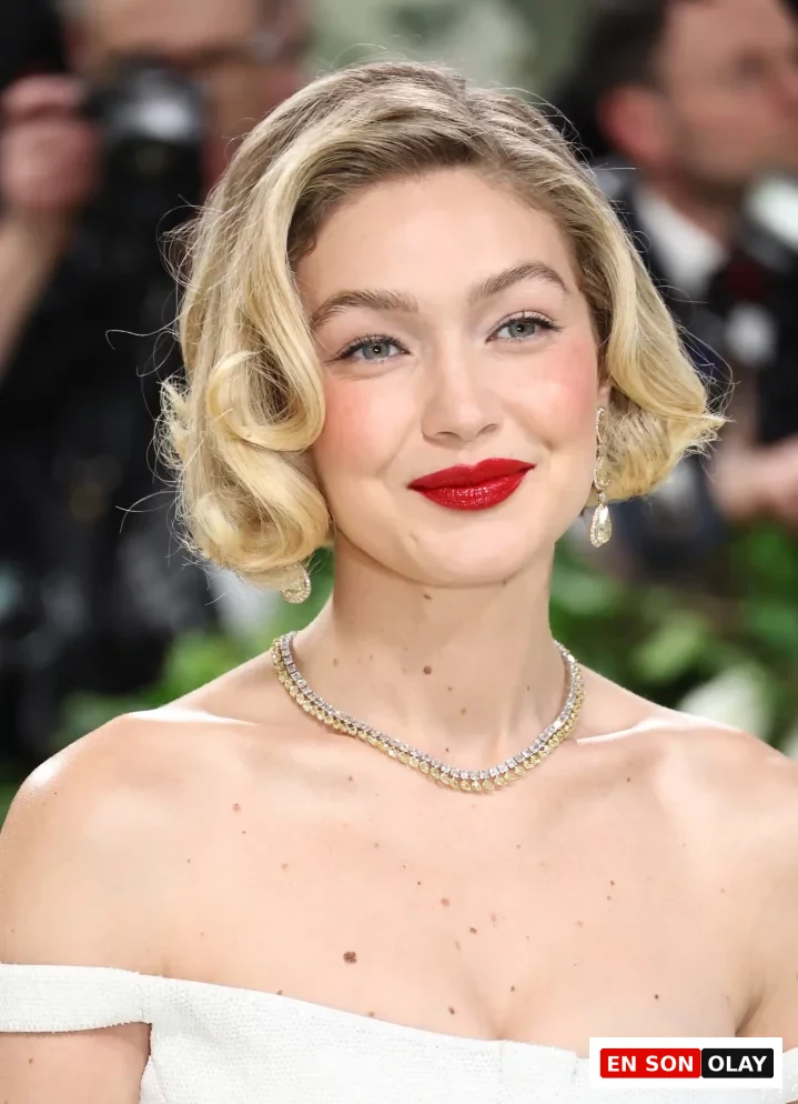 Gigi Hadid, beyaz straplez bir elbiseyle, ikonik dalgalı saç modeliyle zarif bir şekilde gülümserken, dudaklarında parlak kırmızı ruj ve boynunda ışıltılı pırlanta bir kolye ile yakın plandan görünüyor.