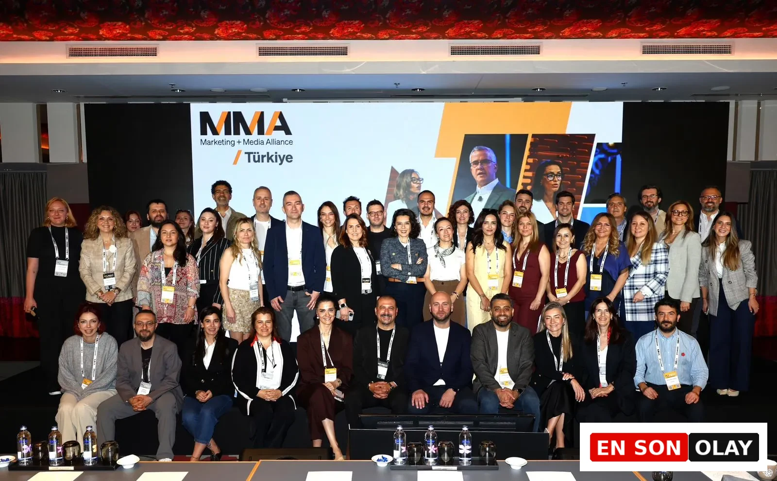 MMA Marketing + Medya İttifakı Türkiye logosunun büyük bir ekranda belirgin şekilde görüldüğü bir konferans salonunda, farklı yaş ve cinsiyetten pazarlama ve medya profesyonelleri, etkinliğin ruhunu yansıtan kalabalık bir grup fotoğrafı için bir araya gelmiş.