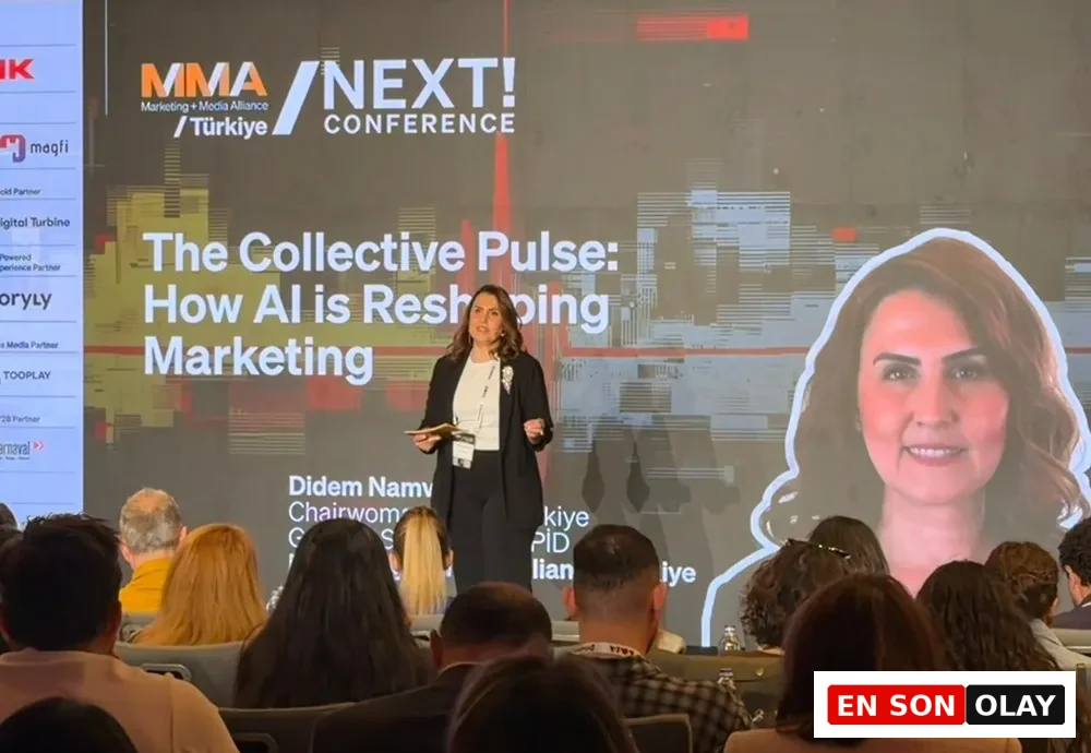 MMA NEXT! Konferansı'nda sahnedeki Didem Namver, büyük bir ekranda Yapay Zeka'nın Pazarlama'yı nasıl yeniden şekillendirdiğini gösteren sunumu yaparken izleyicilere dönük konuşuyor ve ekranda kendisinin gülümseyen bir portresi de görülüyor.
