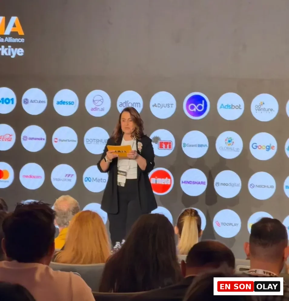 Sahne üzerinde, arkasında birçok şirketin logosu ve MMA Türkiye yazısı bulunan bir fonda, Didem Namver siyah blazer ceketi ve beyaz bluzuyla elinde sarı not kağıtları tutarak dikkatle sunumunu yapıyor, ön planda ise dinleyicilerin başları görülüyor.