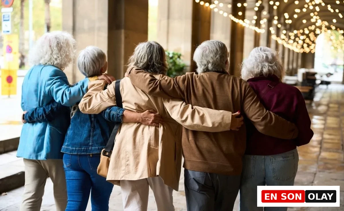 Omuzlarında kolları birbirini saran, arkadan görünen, beyaz saçlı beş yaşlı kadın, ışıklı bir geçidin altında birlikte yürüyerek kadınlar için longevity ve yaşam kalitesi hedefini destekleyen aktif ve güçlü bir sosyal bağı sergiliyor.