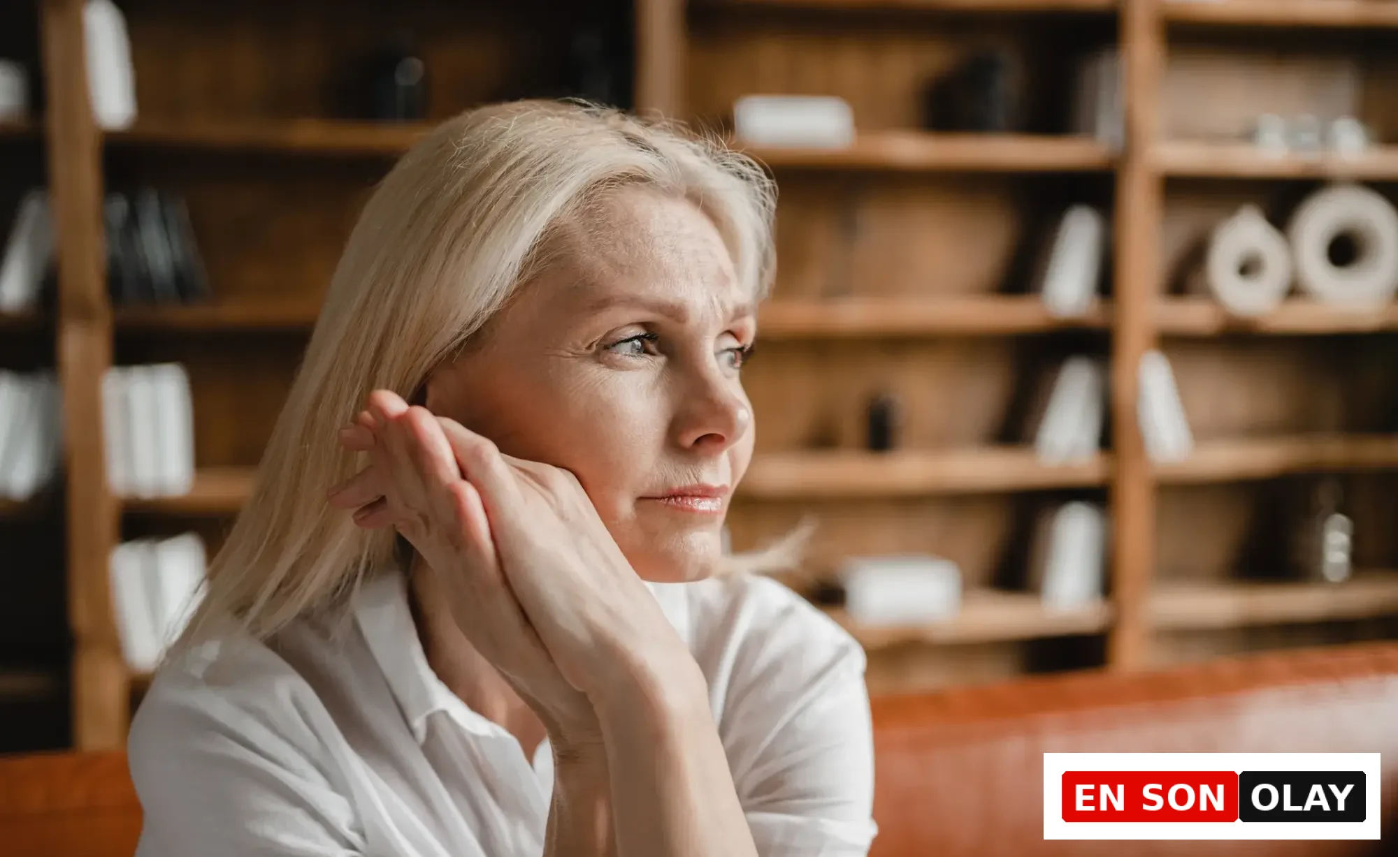 Beyaz gömlekli, sarı saçlı ve düşünceli bakışlara sahip orta yaşlı bir kadın, başkalarının duygusal yükünü taşımanın getirdiği empatik yorgunluğu yansıtan bir ifadeyle, kendi sınırlarını nasıl çizeceğini düşünürken görülüyor.