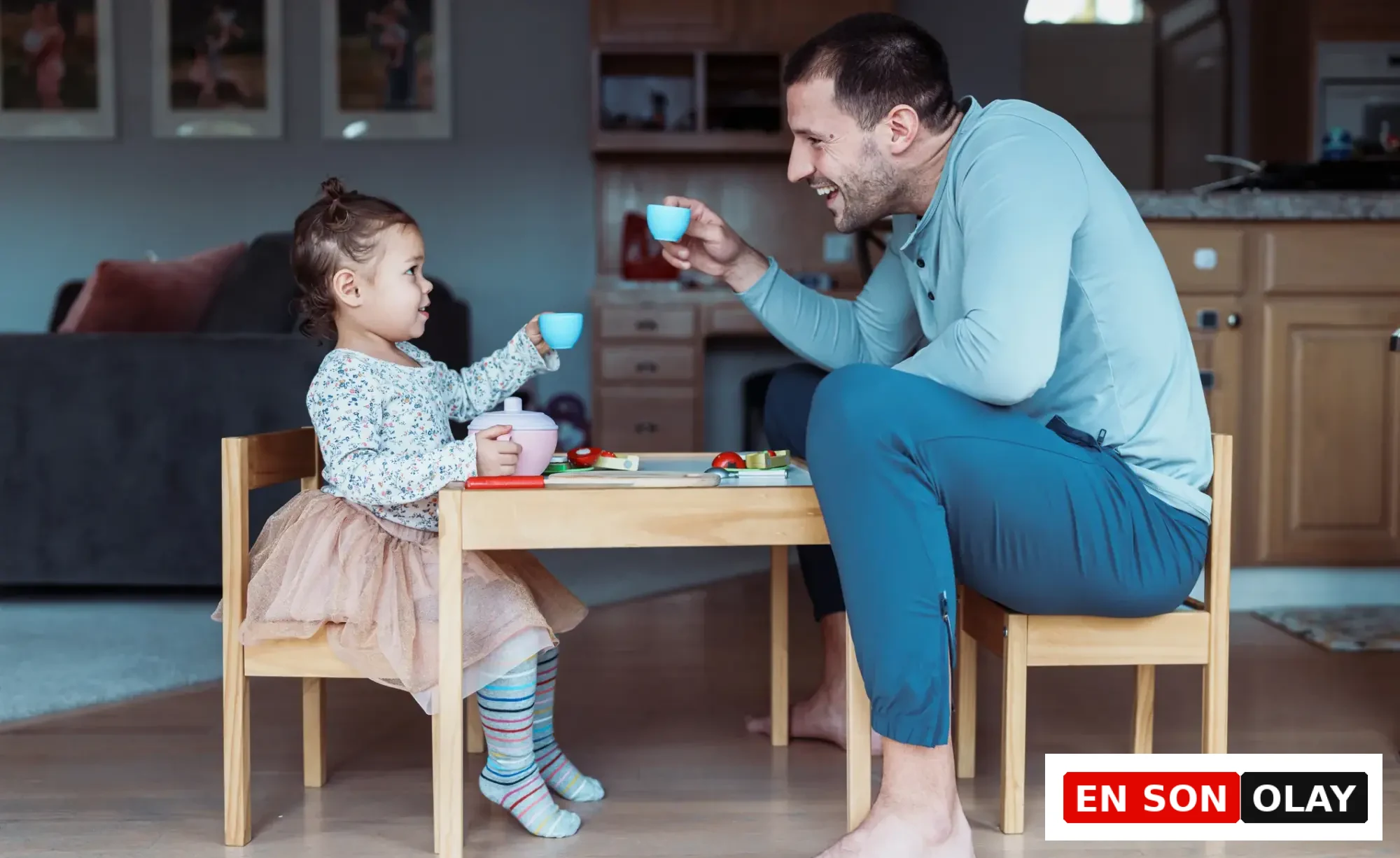 Küçük bir kızın oyuncak masasında, babasıyla karşılıklı oturup oyuncak çay fincanlarını tokuşturarak neşeyle gülümsediği bu içten an, ebeveyn ve çocuk arasındaki güçlü duygusal bağı ve karşılıklı empatiyi hissetmenin en güzel örneklerinden birini sunuyor.