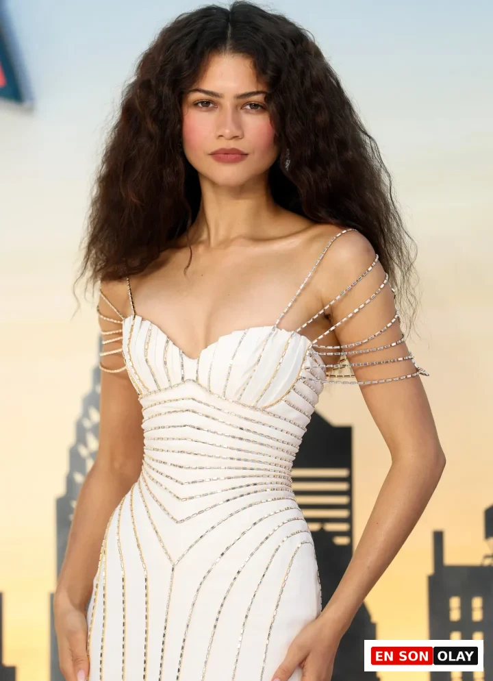 Zendaya, doğal koyu kahverengi, yoğun kıvırcık saç modeli ve zarif makyajıyla öne çıkarken, omuzlarından aşağıya dökülen şık zincir detaylarına ve gümüş ile altın boncuk işlemelerine sahip beyaz bir elbise içinde, sakin bir ifadeyle doğrudan objektife bakıyor.