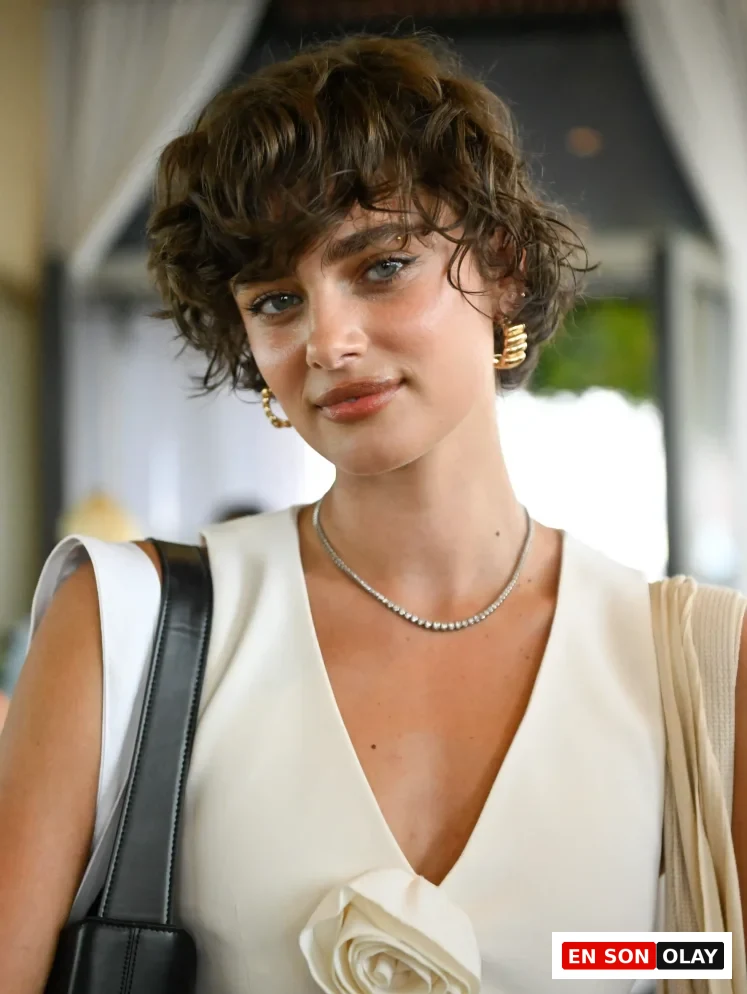Taylor Hill, hafifçe gülümserken, karakteristik kıvırcık saçlarına modern bir hava katan ve alnına düşen belirgin bukleleriyle bixie saç modelini sergiliyor; derin V yakalı krem rengi bluzu, pırlanta kolyesi ve gold renkli halka küpeleriyle zarif ve şık bir görünüm sunuyor.