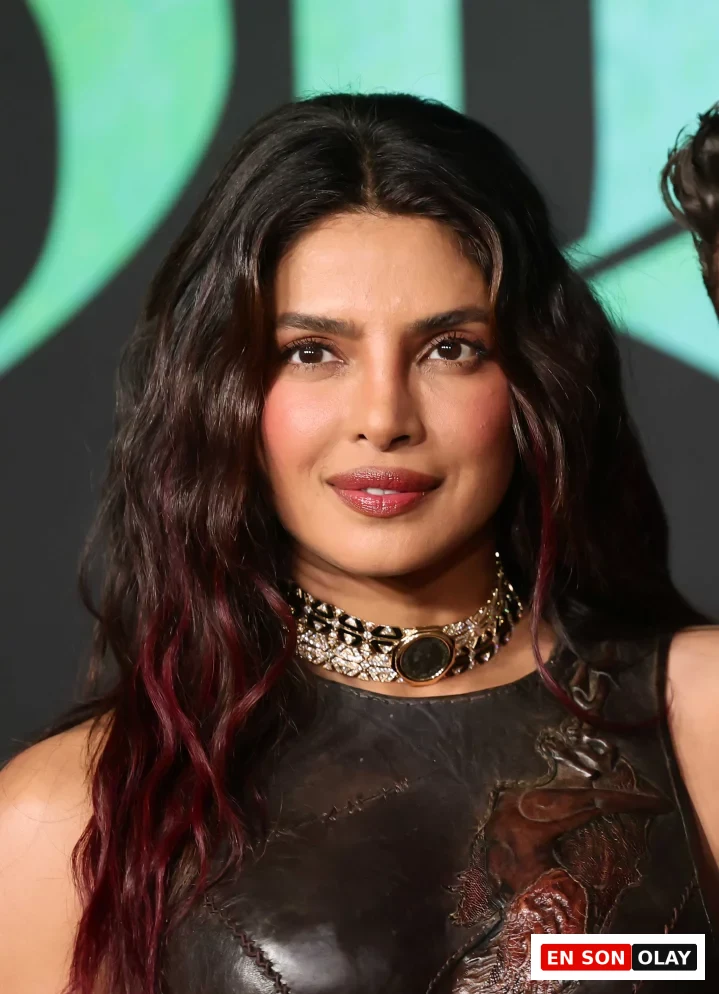 Priyanka Chopra, uçlarında bordo yansımalı koyu dalgalı saçları, göz alıcı işlemeli deri bir üstü ve detaylı taşlı gerdanlığıyla kameraya gülümseyerek bakıyor.