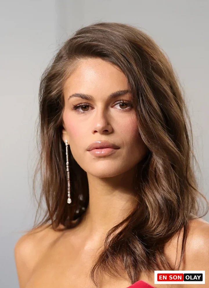 Kaia Gerber'in omuzlarından dökülen uzun, parlak kahverengi ve belirgin dalgalı saç modeli, doğal makyajı, şeffaf dudak parlatıcısı ve göz alıcı uzun pırlanta küpeleriyle kırmızı straplez kıyafetini tamamlayarak zarif bir duruş sergiliyor.