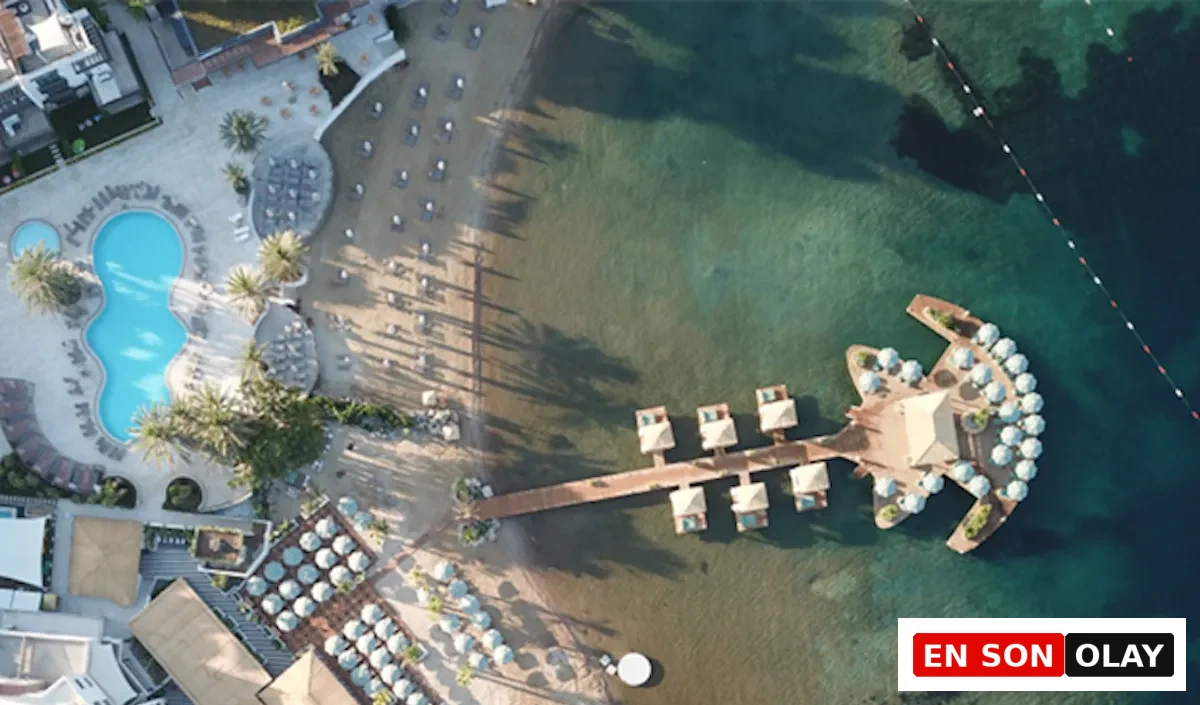 Kuşbakışı görünümde, Bodrum Yalıkavak'ta lüks bir sahil tesisine ait berrak turkuaz deniziyle buluşan kumlu plaj, havuz alanı, ahşap iskele üzerindeki şık su üstü bungalovlar ve iskelenin ucundaki çok sayıda şemsiye ile donatılmış geniş, gitara benzer özel bir platform dikkat çekiyor.