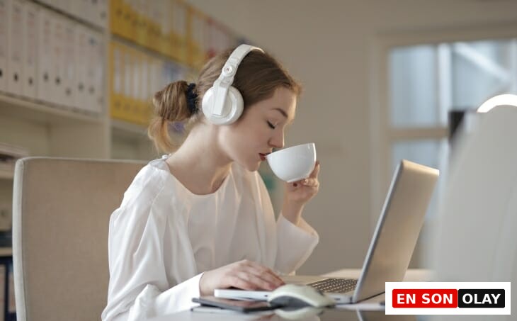Beyaz kulaklığıyla dış dünyadan soyutlanmış, masasında laptop başında yoğun bir şekilde çalışan genç bir kadın, beyaz bir fincandan içeceğini yudumlarken, evden çalışırken verimli olmanın kişisel püf noktalarını hayata geçirdiğini yansıtıyor.