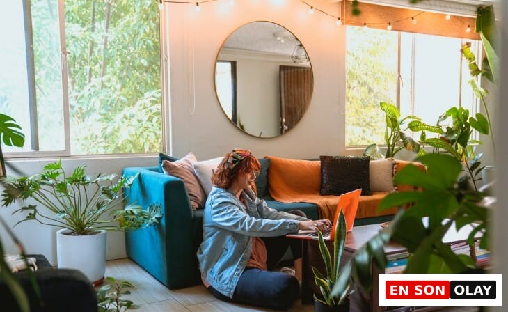 Kırmızımsı kahverengi saçlı, kot ceketli bir kadın, bitkilerle dolu, sıcak bir oturma odasında bağdaş kurarak oturduğu yerde turuncu dizüstü bilgisayarında dikkatle çalışırken, evden çalışırken verimli olmanın odaklanmış bir yolunu sergiliyor.