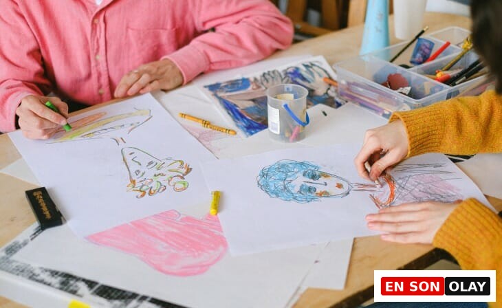 Ahşap bir masada oturan iki kişinin, biri pembe diğeri hardal sarısı kazaklı, farklı renklerde pastel boyalarla yüz çizimleri ve soyut şekillerin olduğu beyaz kağıtları boyarken, evden çalışırken yaratıcılığı ve motivasyonu artıracak mola ve ilham kaynakları bulmanın önemini vurgulayan samimi bir an.