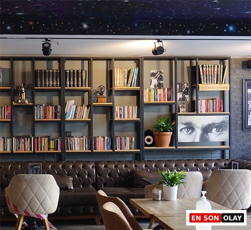 İstanbul'un en iyi çalışma mekânlarından birini gösteren bu iç mekân fotoğrafında, tavanda yıldızlı gökyüzü deseninin altında, arkada kitaplarla dolu metal raflar, saksı bitkileri ve Atatürk'ün dikkat çekici gözlerinin yer aldığı bir tablo öne çıkıyor. Koyu kahverengi deri koltuklar ve ahşap masalarla döşenmiş alanda, ön planda yer alan krem rengi kapitoneli sandalyelerden biri uyarı şeridiyle çevrilmiş, bir masanın üzerinde ise home office çalışanları için düşünülmüş bir el dezenfektanı şişesi duruyor, buranın sakin ve verimli bir çalışma ortamı sunduğunu vurguluyor.