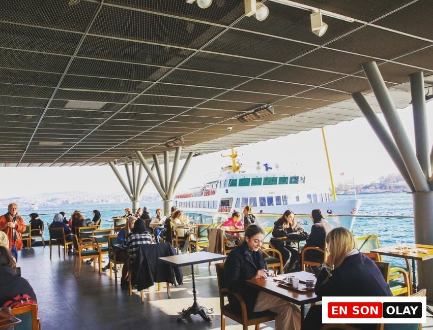 Güneşli bir günde, İstanbul Boğazı'nın eşsiz manzarasına sahip bir teras kafede, insanlar masalarında keyifli vakit geçiriyor; bazıları sohbet ederken, diğerleri sakin bir çalışma ortamı arayanlar için ideal olan bu mekânda işlerine odaklanarak şehrin en iyi çalışma mekânları deneyimini yaşıyor, arka planda bir vapur geçiyor.