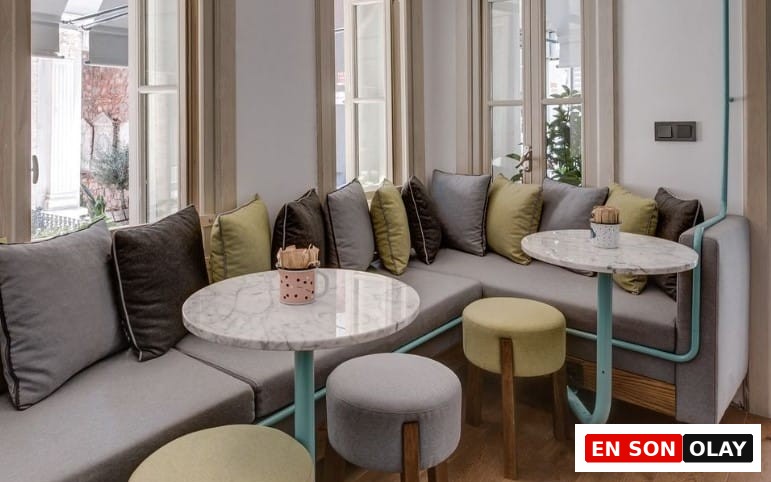 Bir köşeye konumlandırılmış, gri minderli L şeklinde bir kanepeyi çevreleyen, açık mavi metal detaylara sahip yuvarlak mermer masaların, gri ve limon yeşili yastıkların, çeşitli pufların eşlik ettiği bu çağdaş iç mekân, İstanbul'da evden uzakta verimli çalışmak isteyenler için huzurlu ve ilham veren bir çalışma ortamı sunuyor.