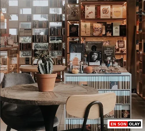 Sıcak bir kitapçı veya kafe ortamında, ortasında saksıda kaktüs bulunan rustik ahşap bir masanın etrafında gri döşemeli ve ahşap sandalyeler yer alırken, arka planda çeşitli kitaplarla dolu ahşap raflar ve dekoratif objelerle zenginleştirilmiş bir kitap sergisi görülüyor.