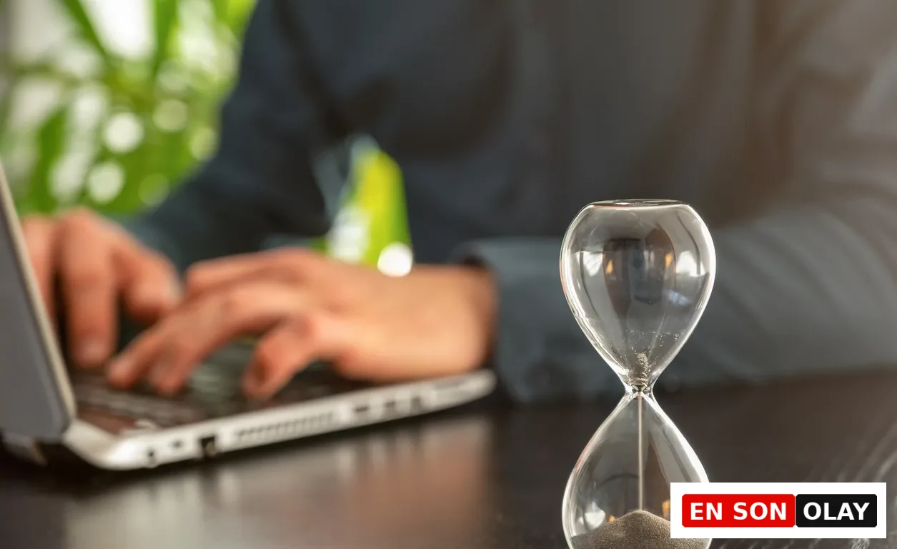 Ensonolay, Festina Lente: Hız Çağında Kontrolü Hatırlatan Antik Felsefe. Ön planda net odaklı bir kum saatinden kumlar usulca akarken, arka planda bulanık bir kişi dizüstü bilgisayarda çalışıyor. Sahne, iş hayatındaki hız baskısına karşı zamanın kontrollü, odaklı ve bilinçli ritmini yansıtıyor.