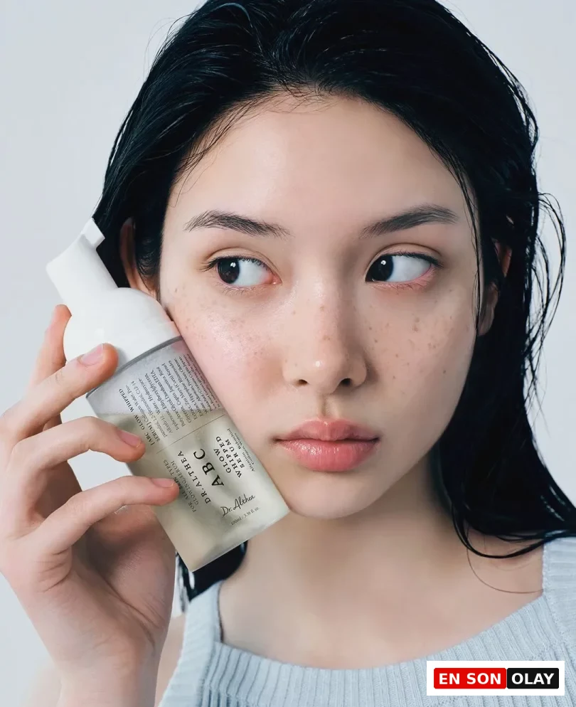 Ortadan ayrılmış ıslak saçları ve çilleri olan genç bir kadın, açık mavi nervürlü bir atlet giyerek Dr. Althea ABC Glow Whipped Serum şişesini hafifçe sol yanağına yaslamış, yan tarafa bakarken Fiddy Metodu ile ilişkili bir cilt bakım ürününü tanıtıyor.