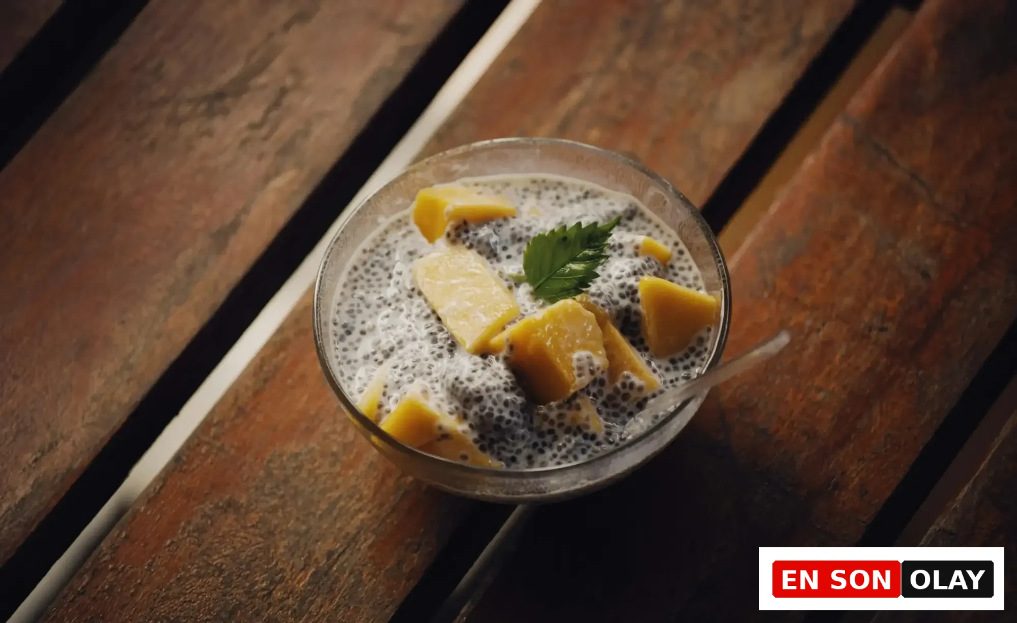 Koyu renk ahşap masa üzerinde, şişmiş chia tohumlarından yapılmış, üzerine canlı sarı mango küpleri ve taze yeşil bir nane yaprağı eklenmiş, sağlıklı ve besleyici bir chia pudingi kasesi görülüyor; bu sunum chia tohumunun çeşitli faydalarından yararlanmak için ideal bir örnektir.