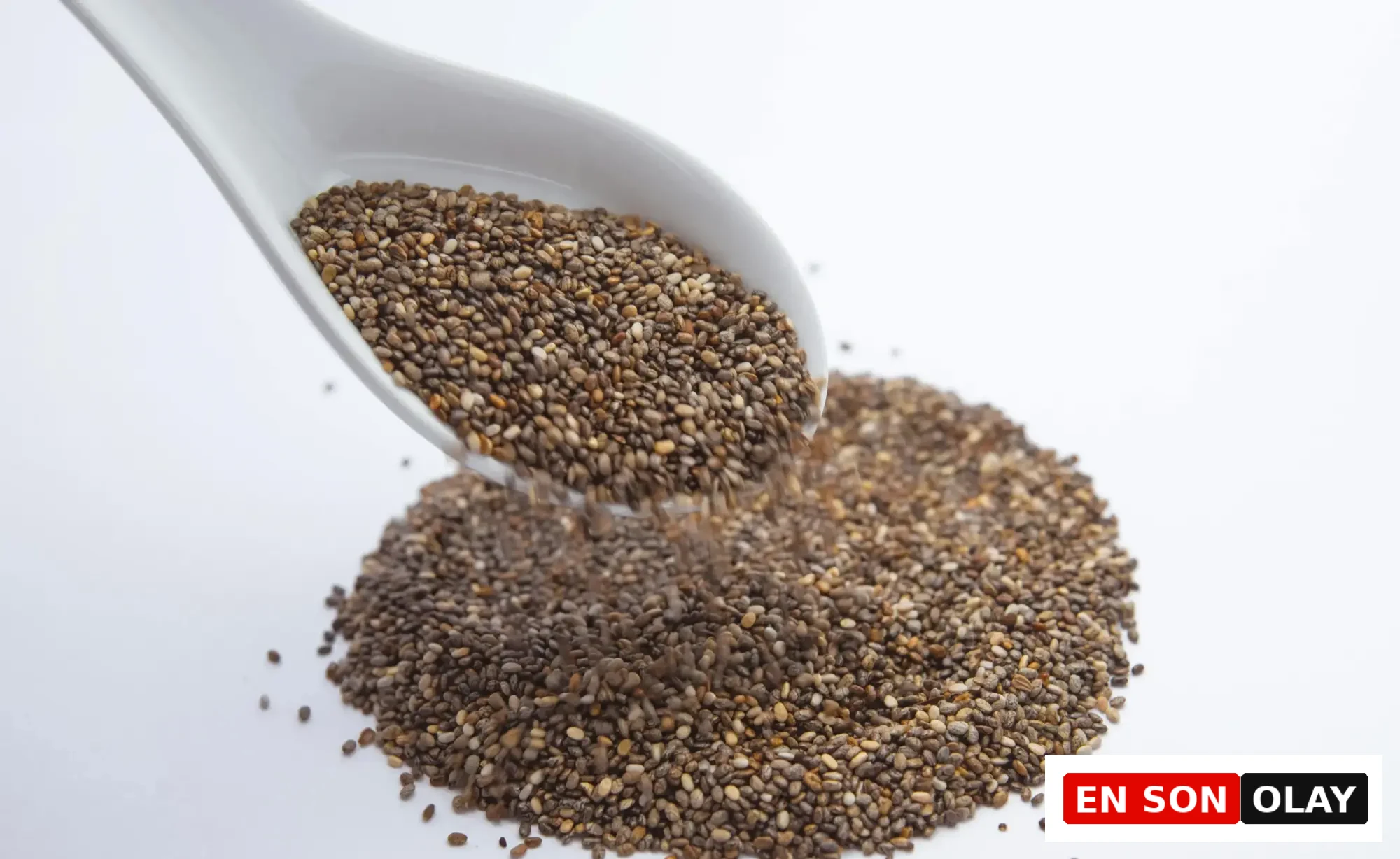 Beyaz bir kepçeden, parlak beyaz bir yüzey üzerinde zaten bulunan yığına doğru dökülmekte olan küçük, kahverengi ve gri tonlardaki sayısız chia tohumu, sunduğu besleyici faydaları ve sağlıklı tariflerdeki kullanım çeşitliliğiyle öne çıkıyor.