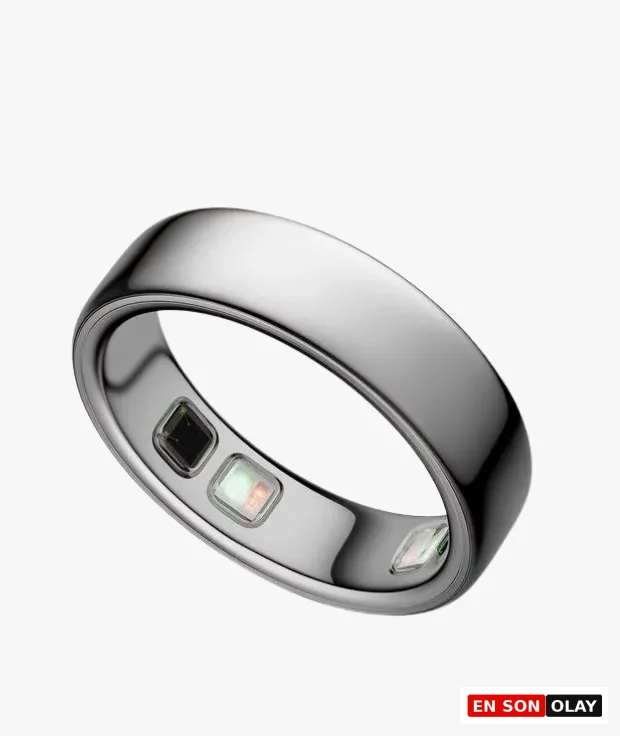 Beyaz bir arka plan üzerinde hafifçe eğik duran, gümüş rengi ve minimalist tasarımlı bir Oura Ring, iç kısmındaki sensörleri belirgin bir şekilde göstererek bu ekransız akıllı wellness yüzüğünün veri toplama özelliğini vurguluyor.