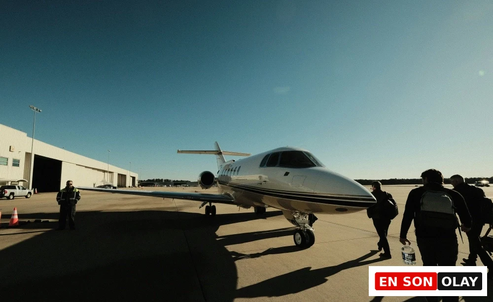 Güneşli açık havada, park etmiş şık bir özel uçağa doğru yürüyen sırt çantalı yolcuların bulunduğu asfalt pist manzarası, sol tarafta bir hangar binası ve yerde bekleyen bir görevli gölgelerle birlikte betimleniyor, konforlu bir yolculuğa çıkış anını yansıtıyor.