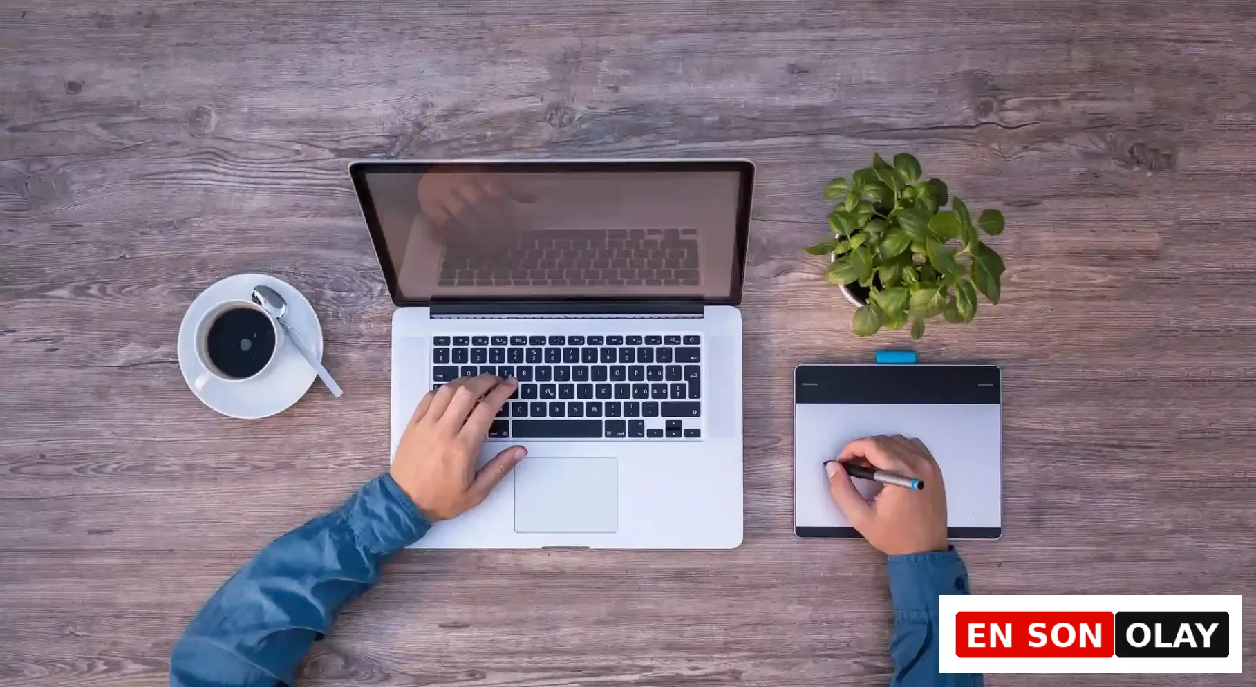 Bir ahşap masa üzerinde, bir yandan dizüstü bilgisayarda yazı yazan diğer yandan grafik tablet kullanan kişinin elleri, ofis ortamında verimli ve odaklı çalışma için kahve ve küçük bir bitki ile çevrili bir çalışma alanını gösteriyor.